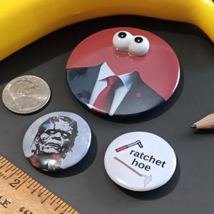 May include: A collection of novelty buttons with various designs. One button features a red background with a suit and tie, topped with googly eyes. Another depicts a monster, and a third has the words "ratchet hoe". A ruler, pencil, and coin are also present.
