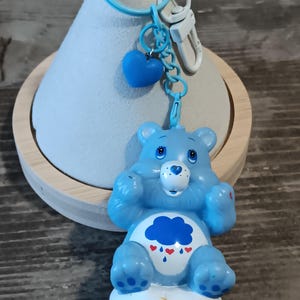 May include: A blue Care Bear keychain featuring a blue bear figure with a cloud and heart design on its belly. The keychain includes a blue heart charm, a blue ring, and a white clasp. The bear is sitting on a white cloud.