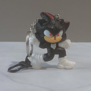 May include: A black and red Shadow the Hedgehog keychain figurine. The figurine is in a crouching pose with white gloves and shoes. The keychain has a silver ring and a black clasp.