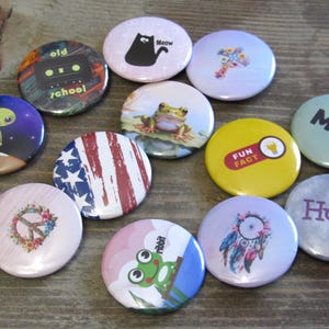 May include: A collection of 12 round button pins with various designs. The pins feature images of a frog, a cat, a peace sign, a dreamcatcher, an alien, a cassette tape, a lightbulb, an American flag, a frog, a cross, and the words "Meh" and "Huh?"