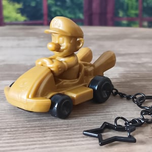 May include: A gold-colored keychain featuring a miniature Mario figure driving a kart. The kart has black wheels and a star-shaped charm attached to the keyring. The figure is wearing a cap with an "M" on it.