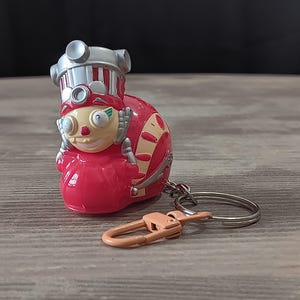 May include: A red and silver cartoon character keychain with a silver keyring and a peach-colored clasp. The character has a round body, a smiling face, and a silver hat. The keychain is on a wooden surface.