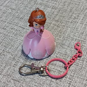 May include: A pink princess figurine keychain with a silver-colored clasp and a pink chain. The princess has brown hair, a tiara, and a pink dress. The keychain is a collectible item.