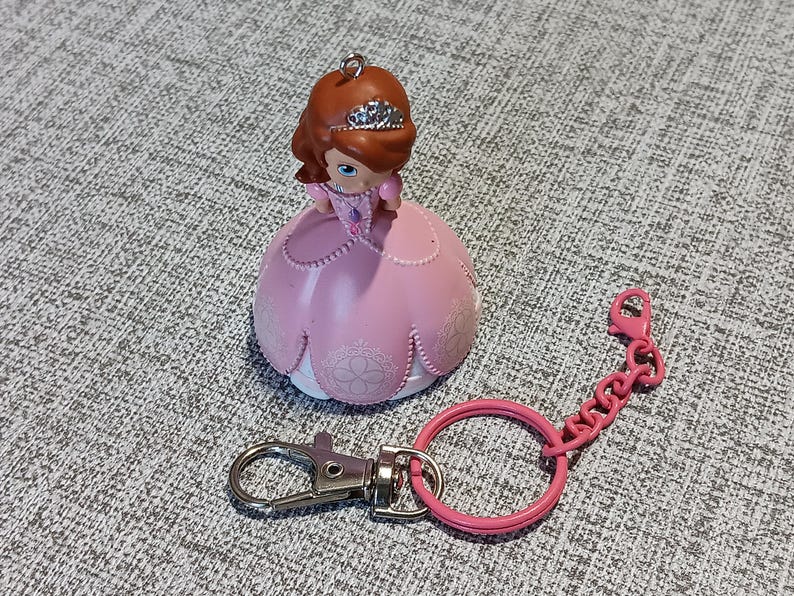 May include: A pink princess figurine keychain with a silver-colored clasp and a pink chain. The princess has brown hair, a tiara, and a pink dress. The keychain is a collectible item.
