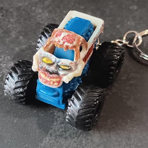 May include: A blue monster truck keychain featuring a white skull-like front grill with yellow eyes and a red brain-like pattern on the roof. The vehicle has four large black tires and a silver metal keyring attached to the rear of the truck body.
