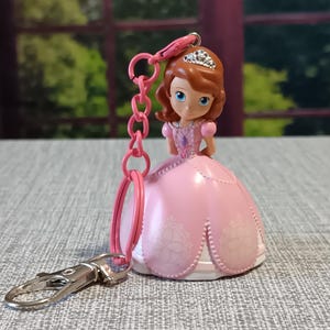 May include: A pink princess keychain featuring a doll with a tiara and a pink dress. The keychain has a silver clasp and a pink chain. The doll has brown hair and blue eyes. The dress has a white floral design.