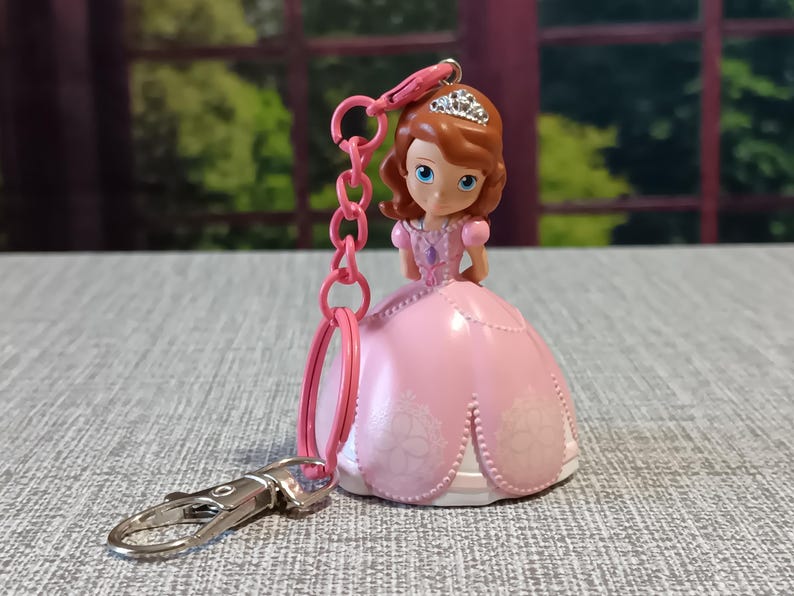 May include: A pink princess keychain featuring a doll with a tiara and a pink dress. The keychain has a silver clasp and a pink chain. The doll has brown hair and blue eyes. The dress has a white floral design.