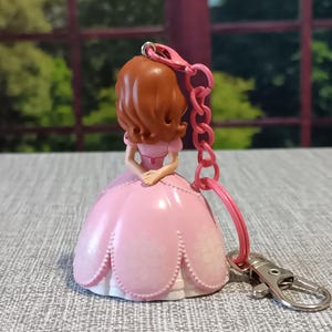 May include: A pink princess keychain with a brown-haired doll wearing a pink dress. The keychain has a pink chain and a silver clasp. The doll's dress has a floral design. The background is blurred, showing green trees.