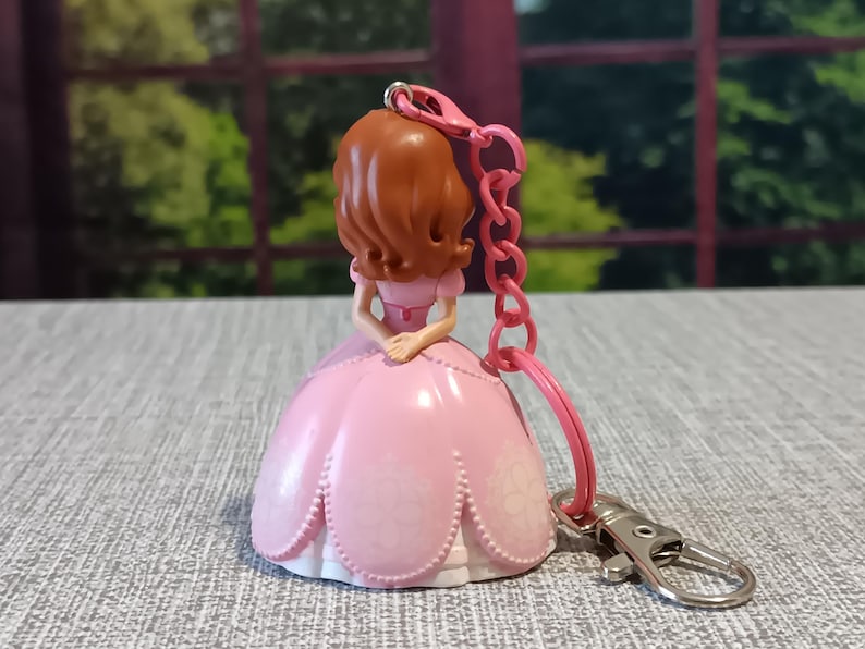 May include: A pink princess keychain with a brown-haired doll wearing a pink dress. The keychain has a pink chain and a silver clasp. The doll's dress has a floral design. The background is blurred, showing green trees.