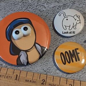 May include: A collection of three round pinback buttons. One button features a cartoon character with large eyes on an orange background. Another button has a white background with a cat illustration and the text "Look at it!". The third button is yellow with the word "OOMF".
