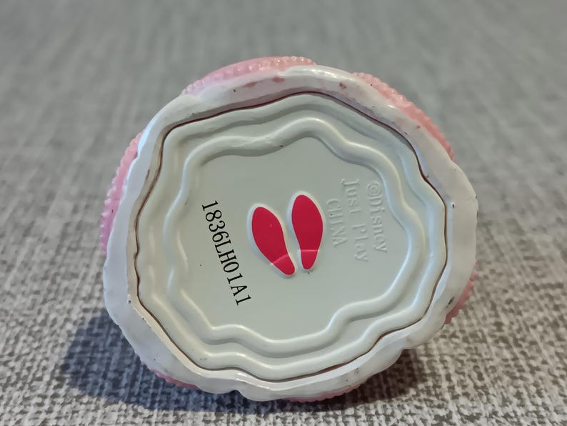 May include: A small, round, pink and white decorative item. The bottom is light gray with the text "&copy;Disney Just Play CHINA" and the number "1836LH01A1". Two red footprints are also visible.