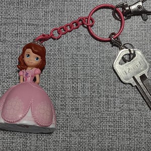 May include: A pink keychain featuring a Sofia the First figurine, a pink chain, a pink ring, and a silver key. The key has the number "12003" printed on it. The keychain is attached to a silver clasp.
