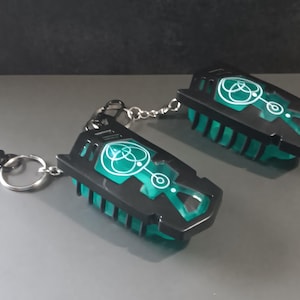 May include: Two black and teal keychains with a geometric design. The keychains have a rectangular shape with a teal interior and a black exterior. Each keychain has a silver chain and a silver keyring. One keychain has a black clip.