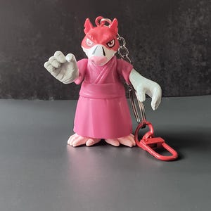 May include: A keychain featuring a stylized anthropomorphic rat figure with a red head, pink snout, and light gray hands. The character wears a solid pink robe with a wrap-style bodice and a matching belt. A silver metal chain and a red plastic clasp are attached.