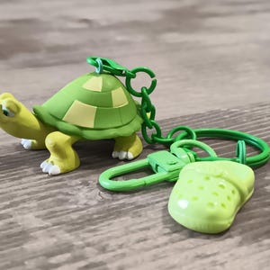 May include: A green and yellow turtle keychain with a matching green shoe charm. The turtle has a green shell with yellow accents and a yellow body. The keychain and charms are attached with green metal hardware.
