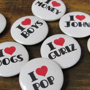 May include: Eight white round buttons with red hearts and black text. The text on the buttons reads "I love", followed by the words "Money", "Boys", "Dogs", "Gurliz", "Pop", "John", "MN", and "N".