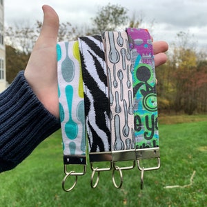 May include: Four colorful fabric keychains with silver key rings. The keychains feature different patterns, including spoons, zebra stripes, and abstract designs. The keychains are held in a hand.