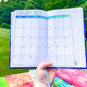 Peut inclure: Agenda ouvert affichant le calendrier de f&eacute;vrier 2023. L'agenda a une couverture sur le th&egrave;me de la galaxie bleue et violette et un ruban bleu clair. Les pages du calendrier sont blanches avec des lignes de grille noires.