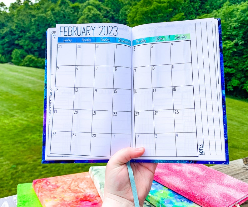 Peut inclure: Agenda ouvert affichant le calendrier de f&eacute;vrier 2023. L'agenda a une couverture sur le th&egrave;me de la galaxie bleue et violette et un ruban bleu clair. Les pages du calendrier sont blanches avec des lignes de grille noires.