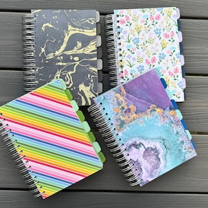 May include: Four different spiral bound notebooks with colorful covers. The covers feature a rainbow stripe pattern, a floral pattern, a marble pattern, and a blue and purple marble pattern.