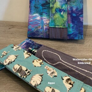 May include: Two fabric pouches, one with a blue and purple watercolour pattern and the other with a light blue background and a white cartoon animal print. The pouch with the watercolour pattern has the text "Watercolor+Grey Embroided" on it. The pouch with the cartoon animal print has the text "Appa" on it. Both pouches have a white embroidered spoon design.