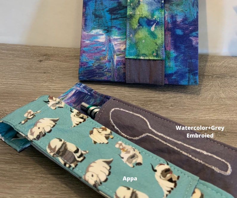 May include: Two fabric pouches, one with a blue and purple watercolour pattern and the other with a light blue background and a white cartoon animal print. The pouch with the watercolour pattern has the text "Watercolor+Grey Embroided" on it. The pouch with the cartoon animal print has the text "Appa" on it. Both pouches have a white embroidered spoon design.
