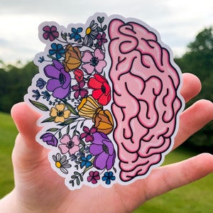 Op de afbeelding: Een roze sticker met een hersenomtrek en een kleurrijk bloemenontwerp. De bloemen zijn in tinten roze, paars, geel en blauw.