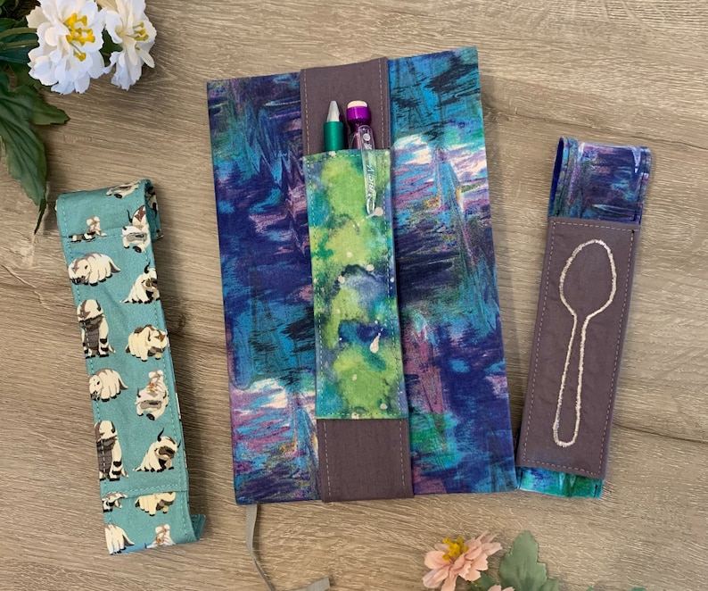 May include: Three fabric pencil holders with different designs. One has a blue and green watercolour pattern, another has a grey background with a white spoon design, and the last one has a teal background with a white pattern of small animals.