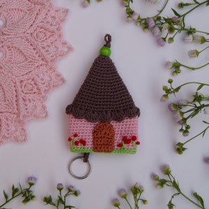 Può includere: Un portachiavi a forma di casa lavorato a uncinetto con una base rosa, un tetto marrone e una porta verde. Il portachiavi ha una piccola perla verde in cima e un anello portachiavi argentato.