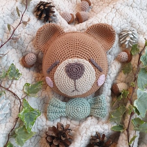Puede incluir: Un oso de crochet marrón con una pajarita verde claro. El oso está sentado sobre una superficie blanca y peluda con pequeñas bellotas marrones esparcidas a su alrededor.