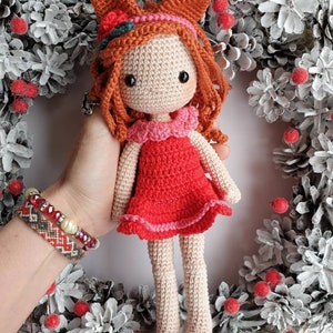 May include: A crocheted doll with red hair, wearing a red dress with pink trim and a pink collar. The doll has a fox-like head with orange ears and is holding a small red berry in its mouth.