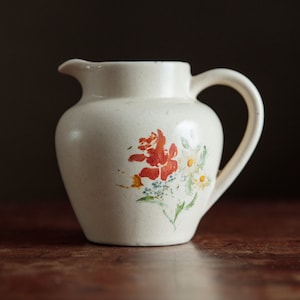 May include: A white ceramic pitcher with a floral design featuring red, orange, yellow, and blue flowers. The pitcher has a handle and a rounded body.