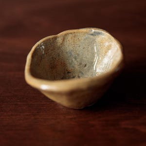Puede incluir: Un pequeño cuenco de cerámica hecho a mano con una forma orgánica única. El cuenco tiene un exterior moteado de color beige y un interior azul claro. El borde es irregular, lo que añade encanto rústico. Se encuentra sobre una superficie de madera marrón oscuro.