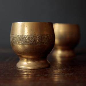May include: Two antique brass goblets with a detailed floral band around the middle. The goblets have a rounded shape and a small footed base. The background is a dark, blurred surface. The goblets are likely used for drinking.