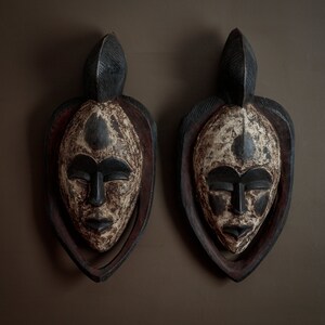 May include: Two carved wooden masks with a tribal design. The masks have a black and white color scheme with a dark brown frame. The masks are mounted on a brown wall.