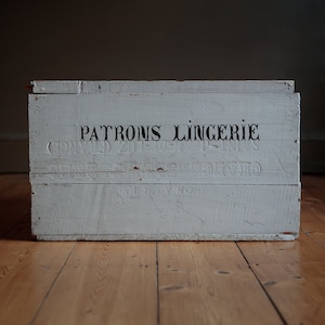 May include: A white wooden box with the text "PATRONS LINGERIE" printed on the front. The box is sitting on a wooden floor.