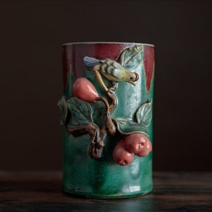 May include: A green ceramic vase with a bird and fruit design. The vase has a red and green glaze and features a bird perched on a branch with red fruit.
