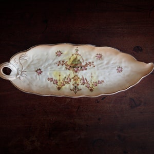 May include: A white ceramic serving dish shaped like a leaf with a floral design in shades of pink, green, and gold. The dish has a raised edge and a handle on one side.