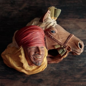 May include: A decorative wall sculpture featuring a man with a red turban and a yellow robe, and a horse with a brown bridle and a white mane. The sculpture is made of painted plaster and has a rustic, vintage look.