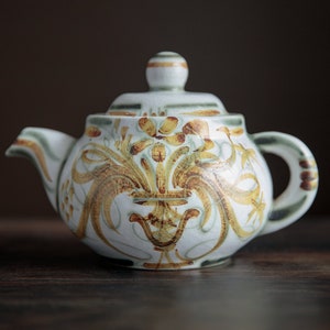 Puede incluir: Una tetera blanca con un diseño floral en tonos marrones y verdes. La tetera tiene un cuerpo redondo y una tapa con un pomo.