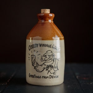 May include: A brown ceramic bottle with a cork stopper. The bottle has a white label with black text that reads "Creedy Vinatge Cider" and "Greetings from Devon". A cartoon illustration of a person holding a glass of cider is on the label.