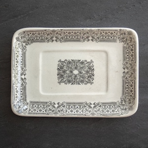 May include: Rectangular off-white soap dish with a black floral and geometric border design. The center features a similar black floral pattern. The dish is made of ceramic and sits on a dark gray surface.