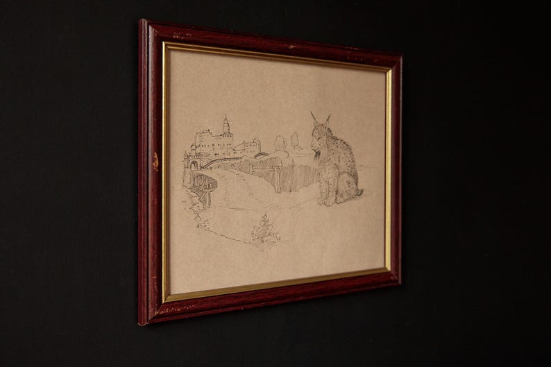 May include: Framed pencil sketch of a lynx and a European castle scene. The artwork is in a dark red wooden frame with a gold inner border. The drawing is on tan paper, and the lynx is seated.