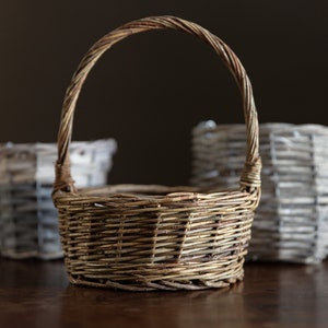 May include: Three woven baskets, one with a handle, on a wooden surface. The basket with the handle is brown, and the other two are gray.