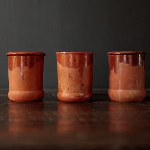 May include: Three terracotta-colored ceramic jars with a glossy, dark brown rim and dripped glaze. The jars are cylindrical with a slightly wider base, set against a dark background. The jars are likely for kitchen or home decor.