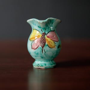 May include: A small, turquoise ceramic vase with a butterfly design. The butterfly has yellow, pink, and brown wings. The vase has a ruffled top and a rounded body, standing approximately 10 cm tall.