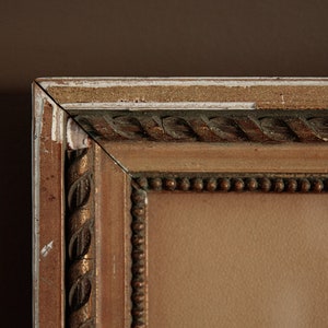Pode incluir: Close-up do canto superior de uma moldura de madeira marrom com detalhes dourados e uma borda de contas. A moldura est&aacute; ligeiramente desgastada com tinta branca aparecendo.