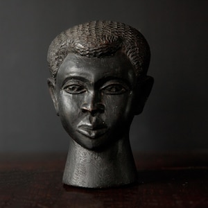 May include: A dark, carved wooden sculpture of a human head. The head is a dark brown colour with detailed facial features, including eyes, a nose, and a mouth. The hair is textured. The sculpture is displayed on a dark wooden surface.