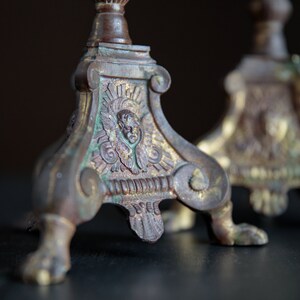 Puede incluir: Un par de candelabros antiguos de lat&oacute;n con detalles ornamentados y una p&aacute;tina verde. Los candelabros tienen un dise&ntilde;o decorativo con una cara en el centro y tres patas.
