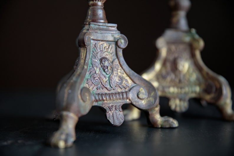 Puede incluir: Un par de candelabros antiguos de lat&oacute;n con detalles ornamentados y una p&aacute;tina verde. Los candelabros tienen un dise&ntilde;o decorativo con una cara en el centro y tres patas.